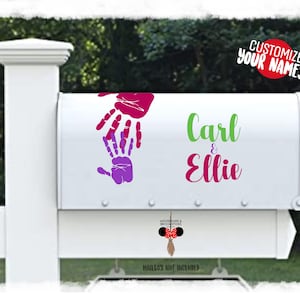 Può includere: Cassetta postale bianca con un design di impronte di mani rosa e viola e i nomi Carl & Ellie scritti in verde e rosa. Il testo "Personalizza i tuoi nomi!" è in rosso.