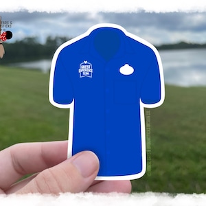 Könnte beinhalten: Ein Aufkleber in Form eines kurzärmeligen blauen Hemdes mit einem weißen Guest Experience Team-Logo auf der rechten Brust und einer weißen, ovalen Namensschildform auf der linken Brust. Das Design umfasst einen Kragen, eine Tasche und einen weißen Rand.