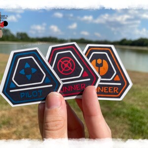 Puede incluir: Tres pegatinas hexagonales con fondo negro y contornos blancos. Las pegatinas tienen centros y símbolos de diferentes colores. La primera pegatina tiene un centro azul con un símbolo de estrella de cuatro puntas y la palabra "PILOT" en blanco. La segunda pegatina tiene un centro rojo con un círculo con una línea a través de él y la palabra "RUNNER" en blanco. La tercera pegatina tiene un centro naranja con un símbolo de llave inglesa y la palabra "ENGINEER" en blanco.