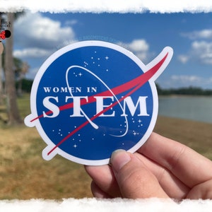 May include: A blue and red sticker with the text "Women in STEM" inside a circle with a red swoosh. The sticker is being held in a hand against a blurred background of a lake and trees.