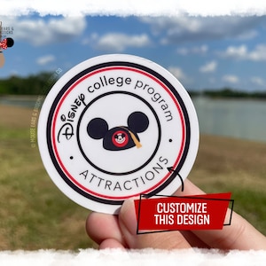 Könnte beinhalten: Ein runder weißer Aufkleber mit rotem und schwarzem Rand. Der Aufkleber hat einen weißen Hintergrund mit dem Text "Disney college program attractions" in einem schwarzen Kreis. Im Kreis befindet sich ein Cartoon-Bild von Mickey Mouse, der einen Abschluss-Hut trägt. Der Aufkleber wird in einer Hand gehalten.