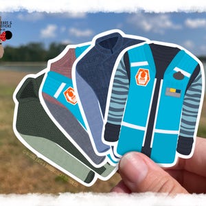 May include: A collection of colorful sticker designs featuring stylized jackets. The jackets are in shades of blue, teal, and green, with various details like stripes and patches. The stickers are held in a hand against a blurred outdoor background.