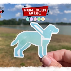 May include: A light blue dog-shaped sticker with wings, held up by a hand. The sticker has a watercolour effect. The text "MULTIPLE COLOURS AVAILABLE" is at the top. The sticker is a design from Mouse Ears & Broomsticks Design Co.