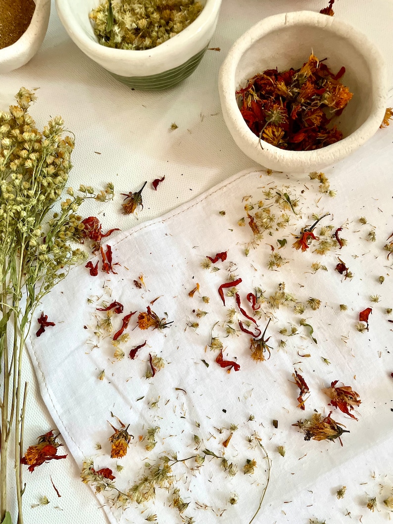 May include: A white linen cloth scattered with dried orange marigold petals and small beige wildflowers. Two small ceramic bowls contain dried herbs, while a bundle of dried goldenrod stems rests to the left of the white fabric.