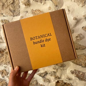 May include: A rectangular brown cardboard box featuring a wide, vertical mustard yellow paper band across the center. The text printed in black on the yellow band reads: BOTANICAL bundle dye kit. The box is held in a hand against a stone wall.