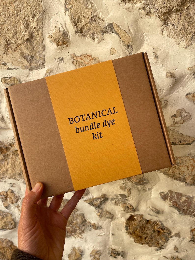 May include: A rectangular brown cardboard box featuring a wide, vertical mustard yellow paper band across the center. The text printed in black on the yellow band reads: BOTANICAL bundle dye kit. The box is held in a hand against a stone wall.
