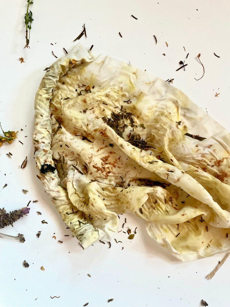 May include: A white fabric pouch with a gathered drawstring closure, stained with yellow and brown botanical pigments. The surface of the cloth is wrinkled and scattered with small, dried green and brown herbal plant fragments.