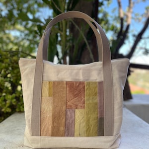 Könnte beinhalten: Eine beige Tragetasche mit braunen Henkeln und einem Patchwork-Design aus verschiedenen Brauntönen, Gelb- und Lilatönen.