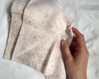 May include: A pale pink fabric square with brown speckles, folded and held in a hand.