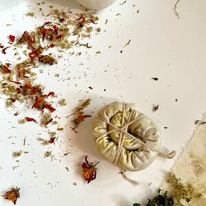May include: A round, fabric bundle tied with thin twine, featuring muted green and yellow botanical dye patterns. Scattered around the bundle are dried orange, yellow, and green flower petals and leaves, along with two small, off-white ceramic bowls.