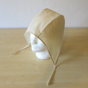 May include: A beige fabric hood with a pointed top and ties. The hood is displayed on a white mannequin head. The fabric appears to be a light cotton or linen, with a simple, clean design. The ties are thin and made of the same material.