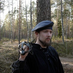 May include: A grey beret and a black long-sleeved shirt with lace-up detailing at the neckline and cuffs. The shirt features a dark, solid fabric, while the beret is a simple, rounded head covering. A metal sword hilt with a cage guard is visible.