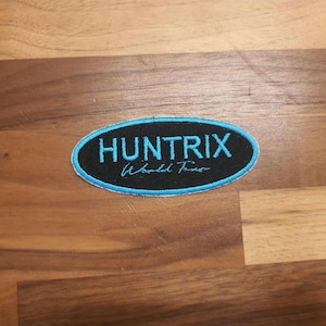 May include: An oval patch with the word "HUNTRIX" in teal, above "World Tour" in a script font. The patch has a black background with a teal border, set against a wood grain surface.
