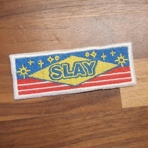 Könnte beinhalten: Gestickter Aufnäher mit dem Wort "SLAY" in Gelb, vor blauem Hintergrund mit Stern- und Diamantakzenten. Der Aufnäher hat ein rot-weiß gestreiftes Design am unteren Rand und einen weißen Rand.
