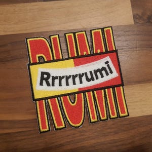 Puede incluir: Parche bordado con la palabra "Rrumi" en un diseño estilizado. El parche presenta una combinación de colores rojo y amarillo con contornos negros y el texto "Rrumi" en blanco.