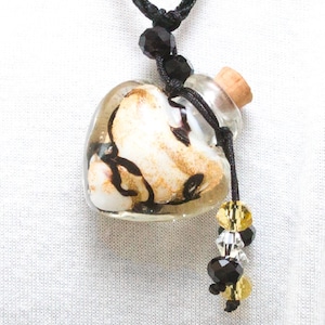 Puede incluir: Un collar con un colgante de botella de vidrio en forma de corazón con un tapón de corcho. La botella está llena de una sustancia blanca y dorada y tiene remolinos negros. El collar tiene cuentas negras y doradas.