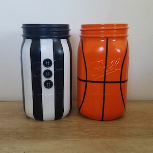 Op de afbeelding: Twee glazen potten geschilderd om eruit te zien als een basketbal en een scheidsrechteruniform. De basketbalpot is oranje met zwarte lijnen en de scheidsrechterpot is zwart-wit gestreept met zwarte knopen.