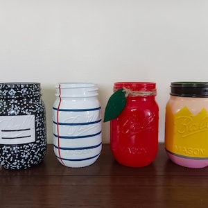 May include: Four painted mason jars. One is black with white speckles, another is white with blue and red stripes, a third is red with a green leaf, and the last is yellow with a pencil design.