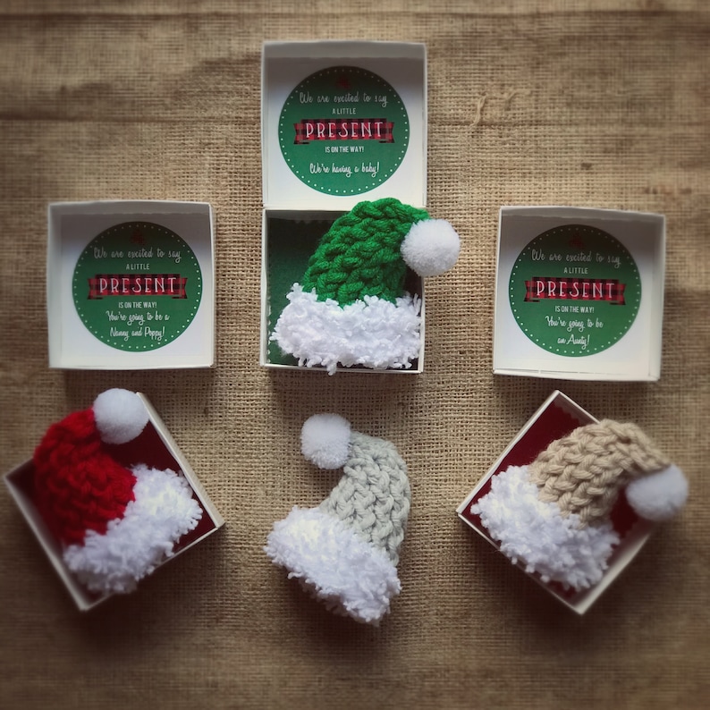 Pu&ograve; includere: Cinque piccole scatole bianche con un cappello di Babbo Natale all'uncinetto all'interno di ciascuna. Le scatole hanno un'etichetta a scacchi rossi e bianchi con il testo "We are excited to say a little present is on the way!"