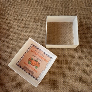 May include: A white gift box with a brown and black checkered pattern on the lid. The lid has a label that says "We are adding a little pumpkin to our patch!" with a drawing of three pumpkins. The label also says "Baby Johnson" and "November 2023".