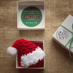 Pu&ograve; includere: Un piccolo cappello di Babbo Natale in crochet rosso e bianco in una scatola bianca con fodera rossa. La scatola &egrave; aperta e il cappello &egrave; visibile. La scatola &egrave; su una superficie di juta marrone. Il testo sul coperchio della scatola dice "We are excited to say a little present is on the way! We're having a baby!"