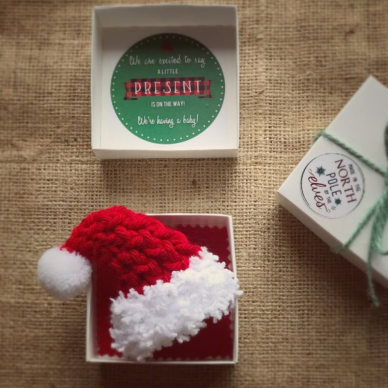 Pu&ograve; includere: Un piccolo cappello di Babbo Natale in crochet rosso e bianco in una scatola bianca con fodera rossa. La scatola &egrave; aperta e il cappello &egrave; visibile. La scatola &egrave; su una superficie di juta marrone. Il testo sul coperchio della scatola dice "We are excited to say a little present is on the way! We're having a baby!"