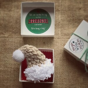 Pu&ograve; includere: Una piccola scatola bianca con un interno rosso contiene un cappello di Babbo Natale di lana bianco e marrone con un pon pon bianco. Il coperchio della scatola ha un cerchio verde con il testo "We are excited to say a little present is on the way! We're having a baby!"