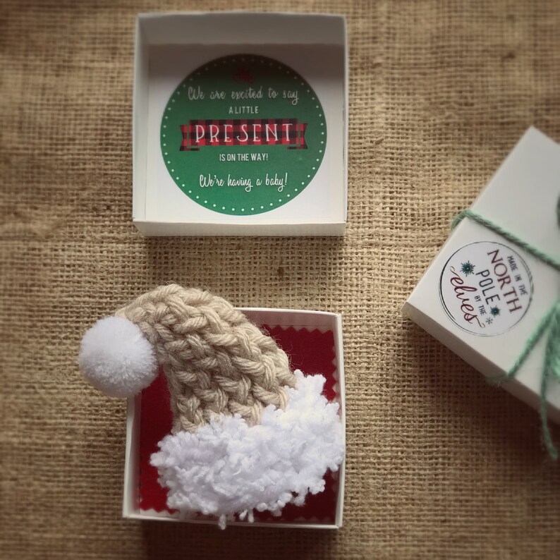 Pu&ograve; includere: Una piccola scatola bianca con un interno rosso contiene un cappello di Babbo Natale di lana bianco e marrone con un pon pon bianco. Il coperchio della scatola ha un cerchio verde con il testo "We are excited to say a little present is on the way! We're having a baby!"