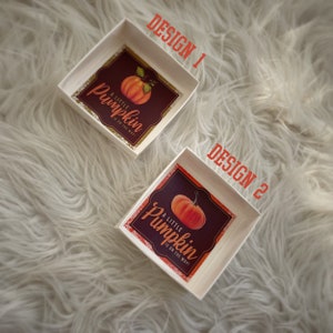 May include: Two white square boxes with a pumpkin design. The boxes have a gold glitter border and the text "A little pumpkin is on the way!" The boxes are on a white fluffy surface.