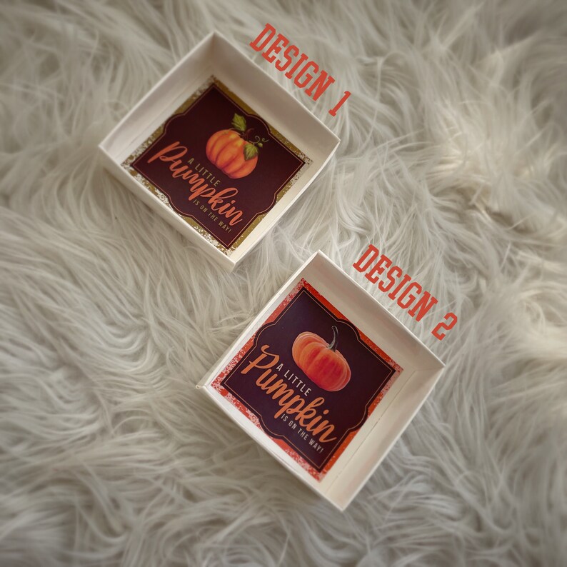 May include: Two white square boxes with a pumpkin design. The boxes have a gold glitter border and the text "A little pumpkin is on the way!" The boxes are on a white fluffy surface.
