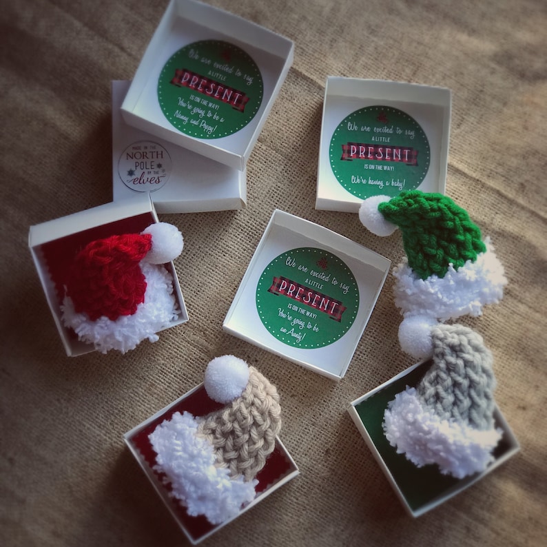 Pu&ograve; includere: Cinque piccole scatole bianche con coperchi, ciascuna contenente un cappello di Babbo Natale all'uncinetto in diversi colori: rosso, verde, grigio e beige. Ogni scatola ha un adesivo rotondo con il testo "Siamo entusiasti di annunciare che un piccolo PRESENTE &egrave; in arrivo! Diventerai una zia!"