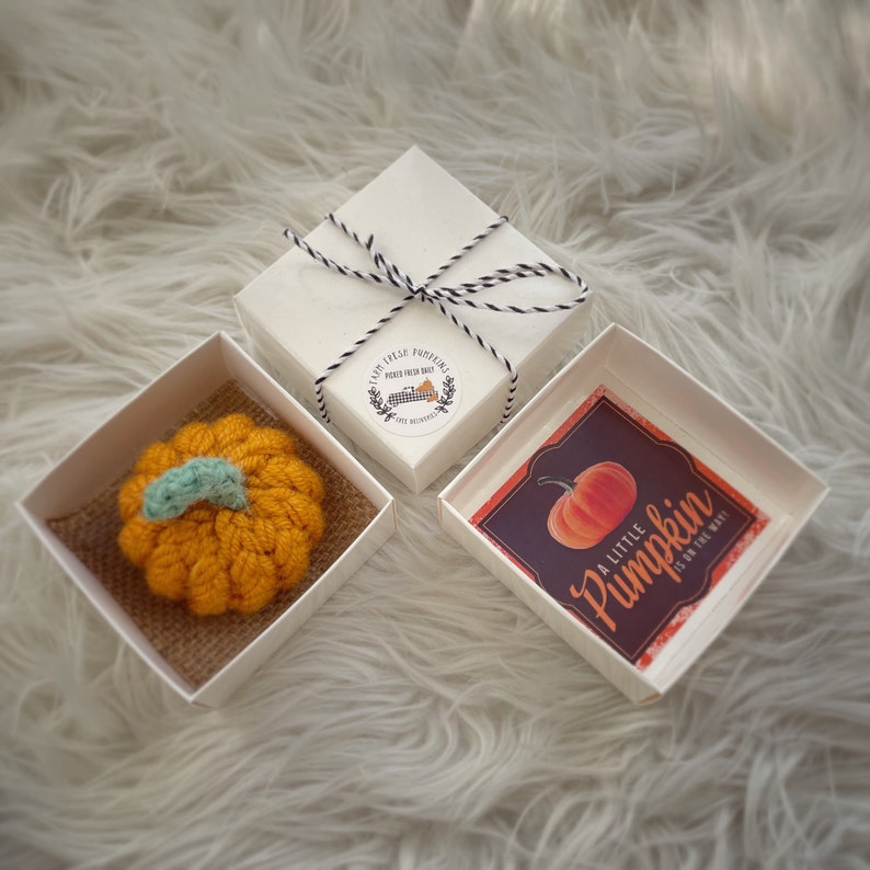 May include: Two white gift boxes with lids. One box contains a small crocheted orange pumpkin with a green stem. The other box contains a card with a red pumpkin illustration and the text "A little Pumpkin is on the way!"
