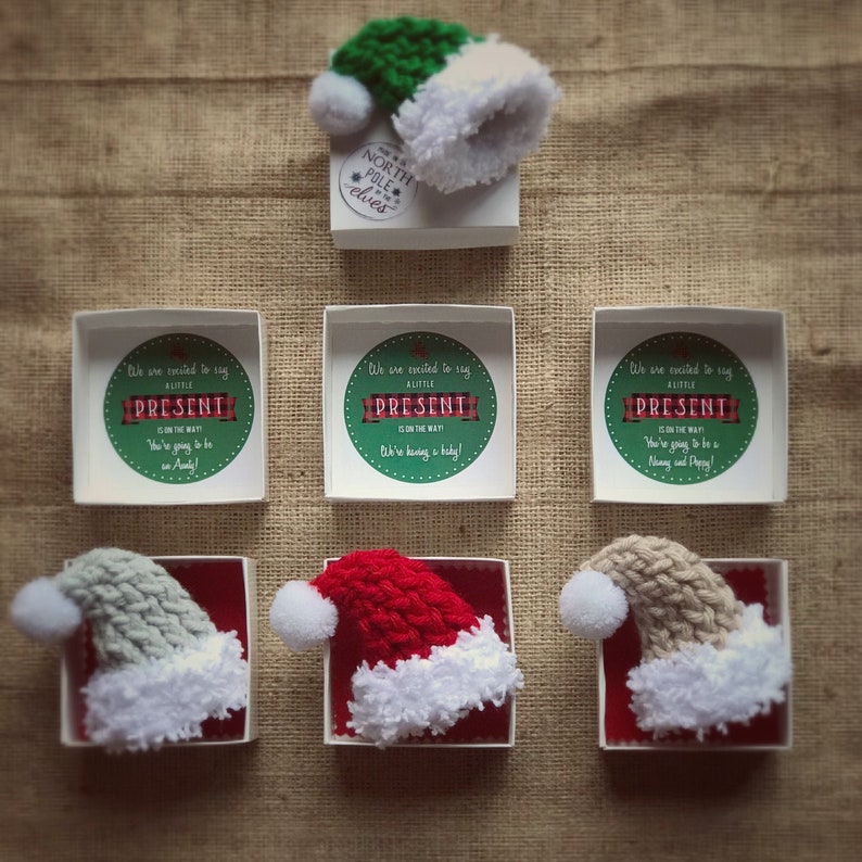 Pu&ograve; includere: Tre scatole bianche con bordi rossi, ciascuna contenente un cappello di Babbo Natale all'uncinetto in diversi colori: grigio, rosso e beige. Ogni scatola contiene un biglietto stampato all'interno che dice "Siamo entusiasti di annunciare che un piccolo regalo &egrave; in arrivo!" con uno sfondo a quadri verde e rosso.