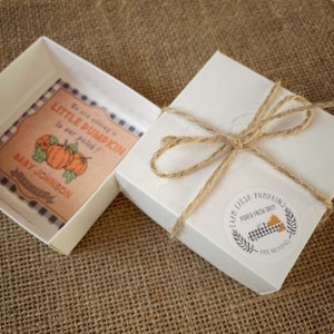 May include: A white gift box with a brown twine bow and a round sticker that says "Farm Fresh Pumpkins Picked Fresh Daily Free Deliveries". The box is open and inside is a small brown card with a black and white checkered border. The card says "We are adding a little pumpkin to our patch! Baby Johnson". There are three orange pumpkins on the card.