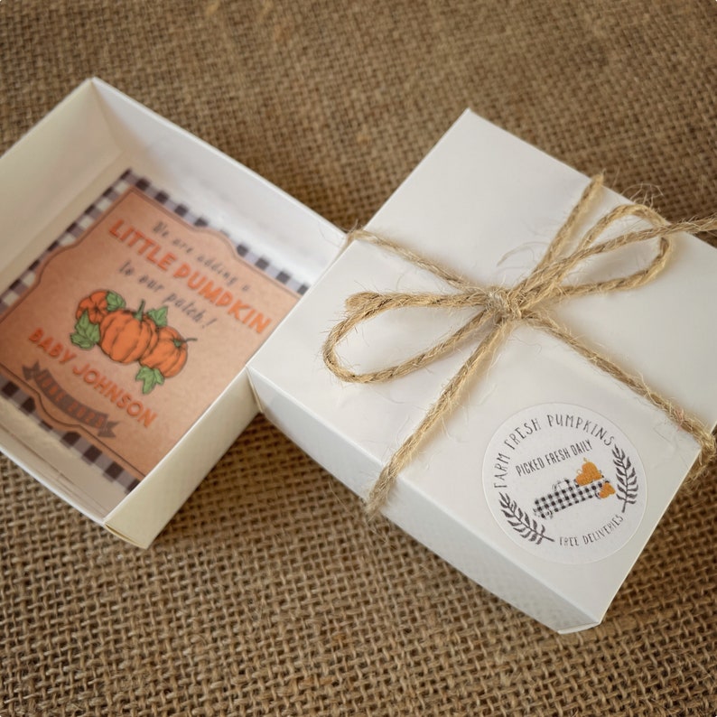 May include: A white gift box with a brown twine bow and a round sticker that says "Farm Fresh Pumpkins Picked Fresh Daily Free Deliveries". The box is open and inside is a small brown card with a black and white checkered border. The card says "We are adding a little pumpkin to our patch! Baby Johnson". There are three orange pumpkins on the card.