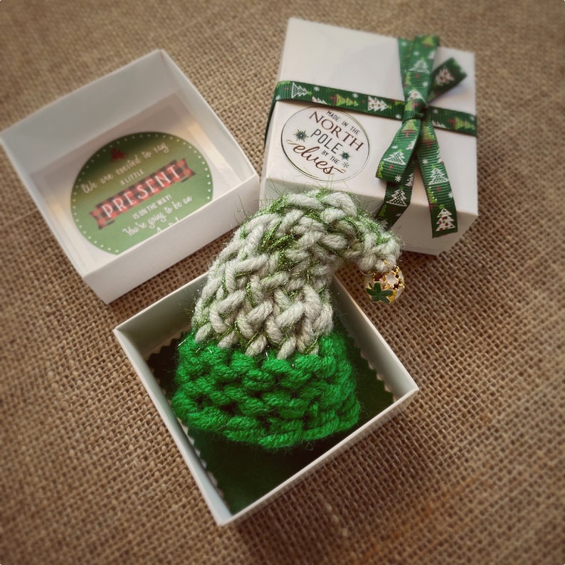 Pu&ograve; includere: Un piccolo cappello da elfo di lana verde e bianco con un ciondolo a campana dorata, confezionato in una scatola regalo bianca con un nastro verde e bianco. La scatola ha un adesivo che dice "Fatto al Polo Nord dagli elfi".