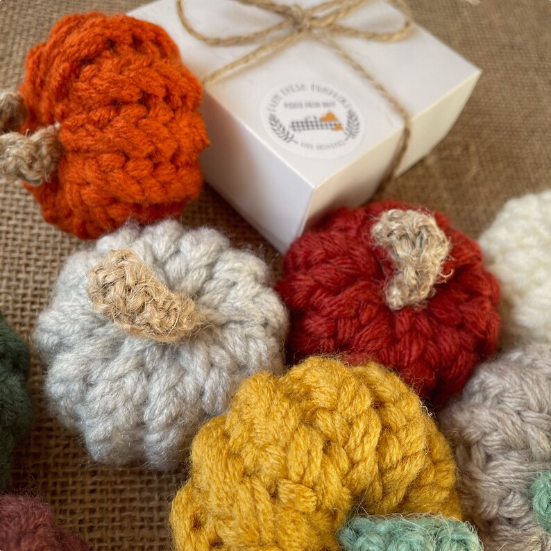 May include: A collection of small crocheted pumpkins in various colors, including orange, red, gray, yellow, and green. The pumpkins are arranged on a burlap surface, with a white box in the background.