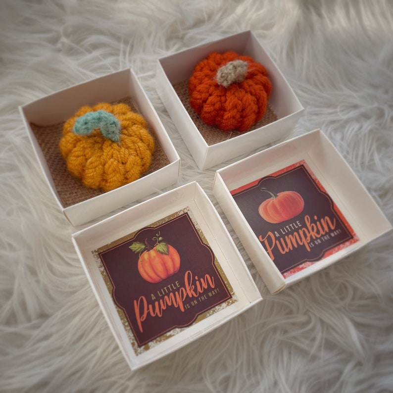 May include: Four small white boxes with lids. Each box contains a crocheted orange pumpkin and a printed card with the text "A little pumpkin is on the way".