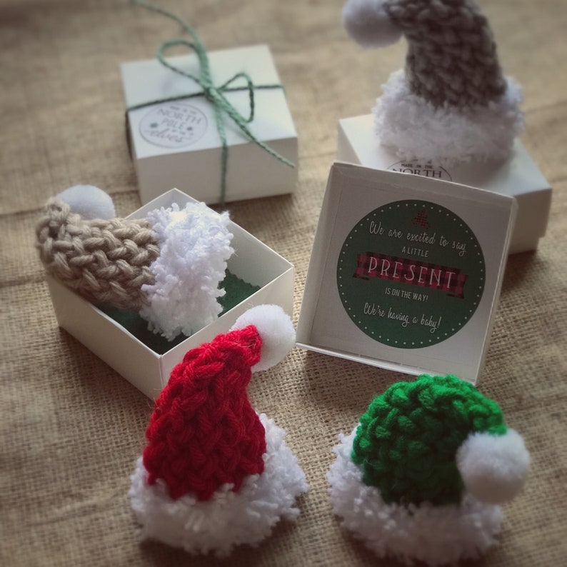 Pu&ograve; includere: Quattro piccoli cappelli di Babbo Natale a maglia in rosso, verde, beige e grigio con pon pon bianchi. I cappelli sono in piccole scatole con il testo "Siamo entusiasti di annunciare che un piccolo regalo &egrave; in arrivo! Stiamo aspettando un bambino!"