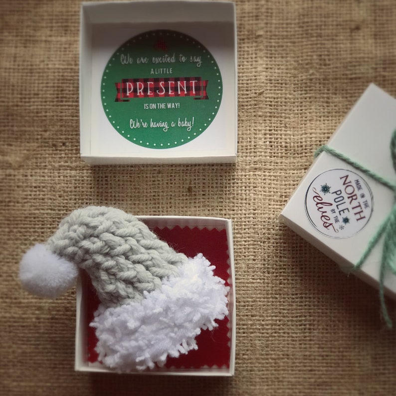 Pu&ograve; includere: Due scatole regalo bianche con accenti rossi e verdi. Una scatola contiene un cappello di Babbo Natale di lana grigia con un pon pon bianco e un bordo di pelliccia. L'altra scatola ha un adesivo che dice "Fatto al Polo Nord dagli elfi".
