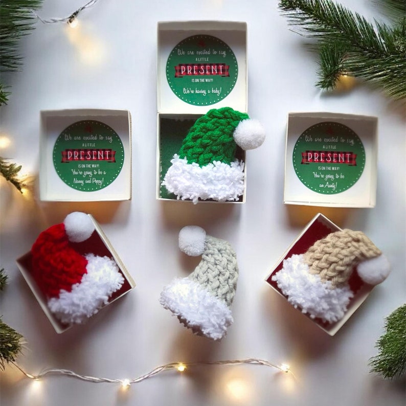 Pu&ograve; includere: Quattro piccoli cappelli di Babbo Natale all'uncinetto in diversi colori, tra cui rosso, verde, grigio e beige, sono esposti in scatole aperte con un annuncio natalizio all'interno di ogni scatola. L'annuncio recita: "Siamo entusiasti di annunciare che un piccolo regalo &egrave; in arrivo!"