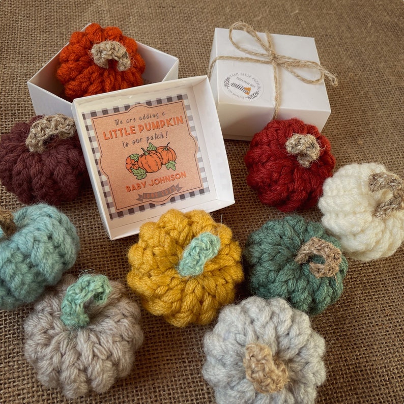 May include: A white gift box with a crocheted pumpkin inside. The box has a label that says "We are adding a little pumpkin to our patch! Baby Johnson" and a small tag that says "Little Feet Boutique". There are other crocheted pumpkins in various colors around the box.