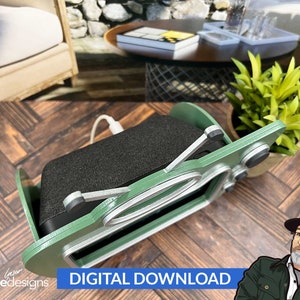 May include: A green and silver wooden stand designed to hold a smart speaker. The stand has a retro design with a circular cutout and a small shelf for the speaker's power cord. The stand is sitting on a wooden surface.