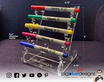 May include: A clear acrylic marker holder with five slots for markers. The holder is 10.75 inches wide, 6 inches tall, and 4.75 inches deep. The slots are 0.6 inches in diameter. The holder is labelled "bigbluedesigns" and has a laser-cut design. The holder is displayed on a blue fabric surface.