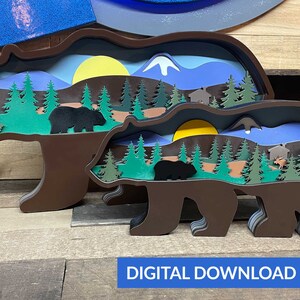 May include: Two brown wooden bear silhouettes with layered cutouts of a forest scene, mountains, and a sun. The larger bear is in front of the smaller bear. The scene inside the bears is a layered cutout of a forest with trees, a sun, mountains, and a small cabin.