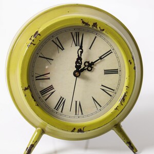 May include: A round table clock with a distressed lime green metal frame and two angled legs. The clock face is off-white with black Roman numerals and ornate black metal hands. The paint on the frame shows signs of wear and dark brown chipping.