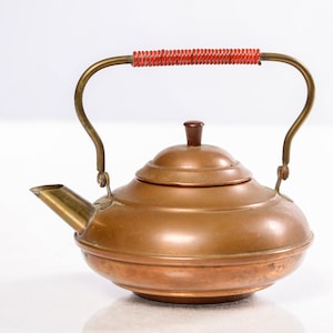 May include: A copper teapot with a brass handle and spout. The handle has a red coiled wrap.