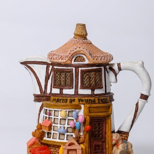 May include: A decorative teapot shaped like a toy shop with a thatched roof, white walls, and brown timber framing. The storefront features the text Merry go round Toys, a brown door, and miniature toys including a red car and a rocking horse.