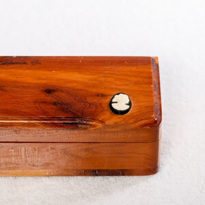 May include: A wooden box with a hinged lid. The box is made of cedar wood and has a small, white cameo on the top. The box is labeled "Lake Junaluska, Ga."