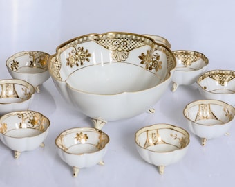 May include: A large central bowl and seven smaller matching bowls, all white with scalloped edges and gold-painted floral and lattice patterns. Each piece features three small feet at the base, creating a cohesive set of decorative serving dishes.